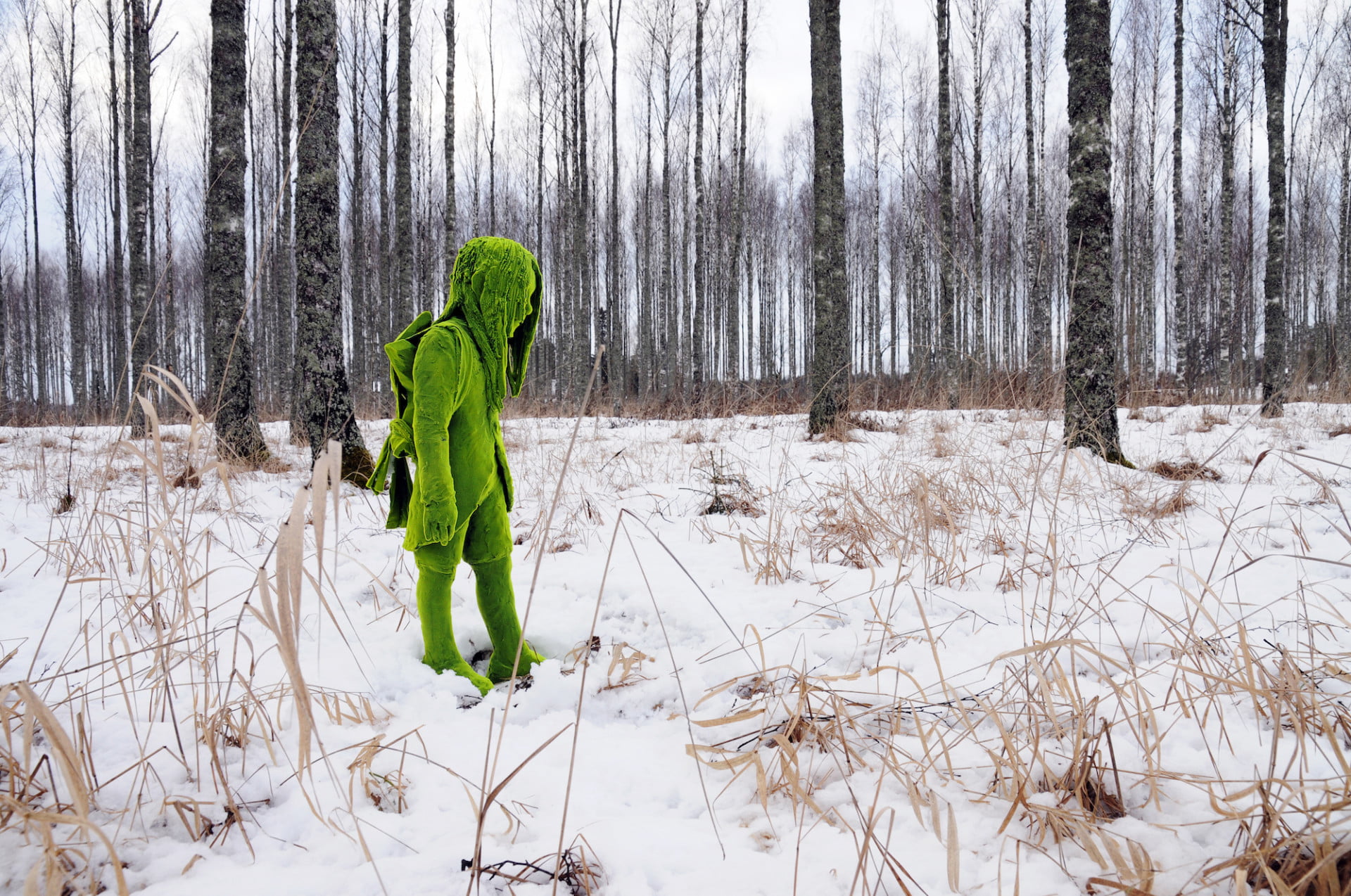 Uma fotografia num bosque nevado de uma escultura cerâmica que foi revestida em flocos verdes para fazer parecer que está revestida de musgo. A escultura é de uma figura jovem de perfil que parece caminhar através da neve.