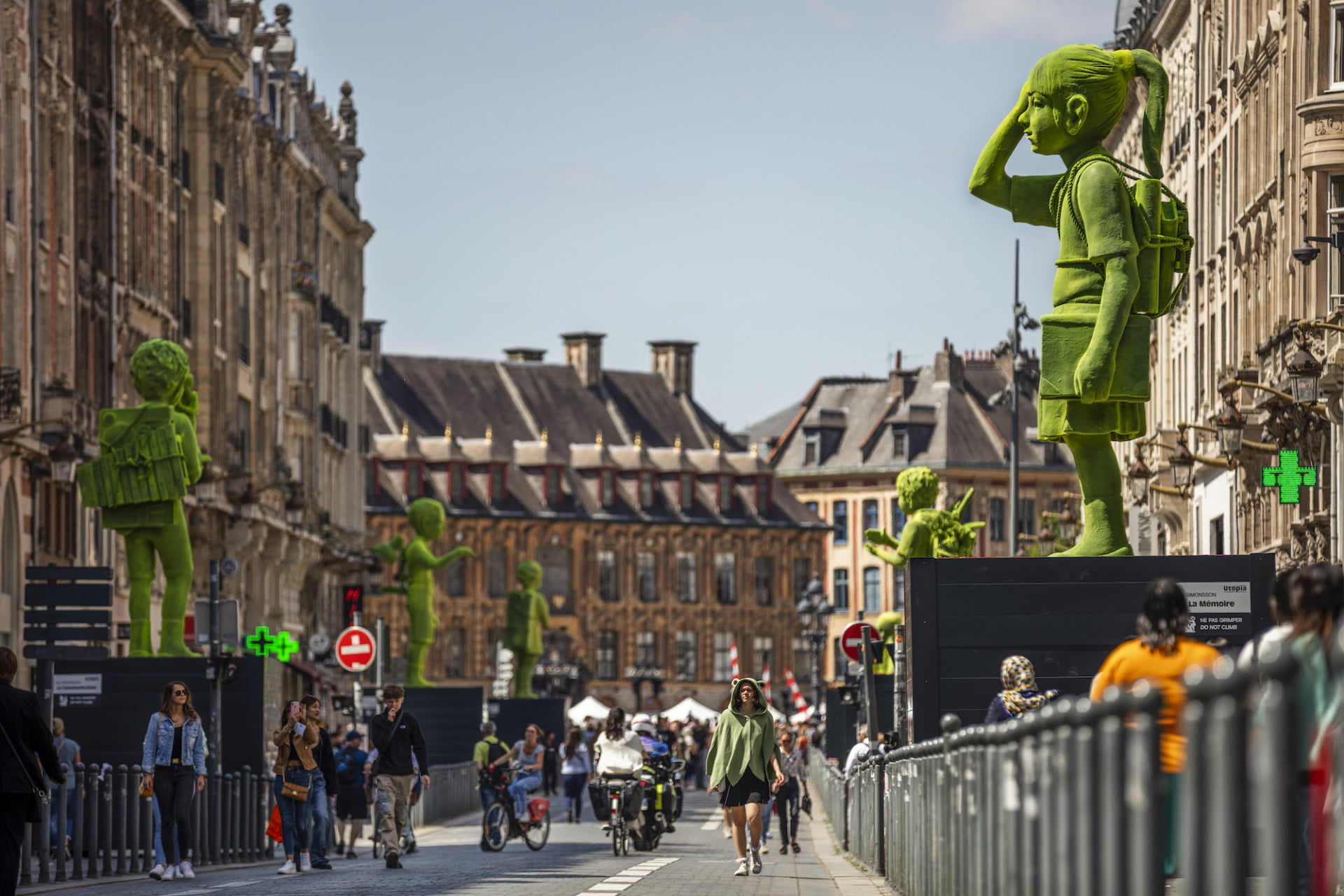 Large-scale fiberglass figurines in a street in Lille, France, retratando figuras que são revestidas em flocos verdes para fazê-las aparecer como se estivessem revestidas de musgo. Os peões descem a rua e as esculturas são exibidas em grandes pedestais.
